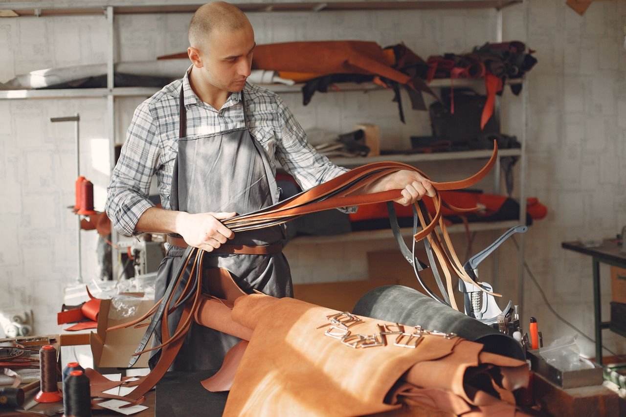 Serious young man with shaved head in apron standing near table with leather and checking belts while working in modern workshop